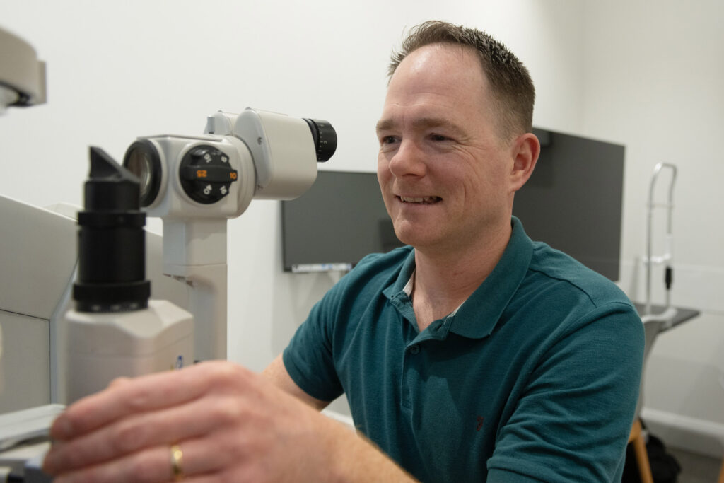 Profile image for Nicholas Sage, Nick is smiling while looking through equipment at a patients eye